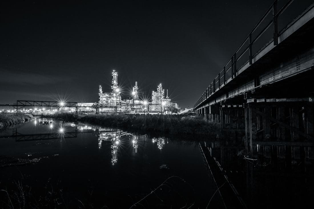 Black and white night photograph of an illuminated refinery reflected in water